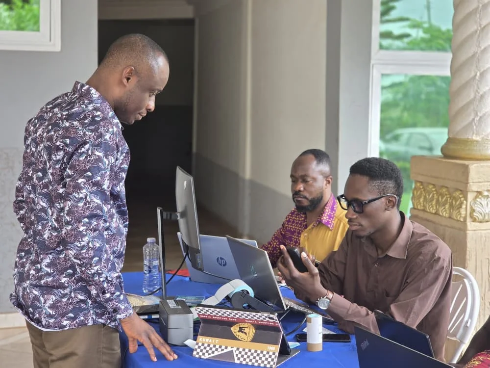 An attendee is assisted at a check-in desk equipped with laptops and a badge printer.